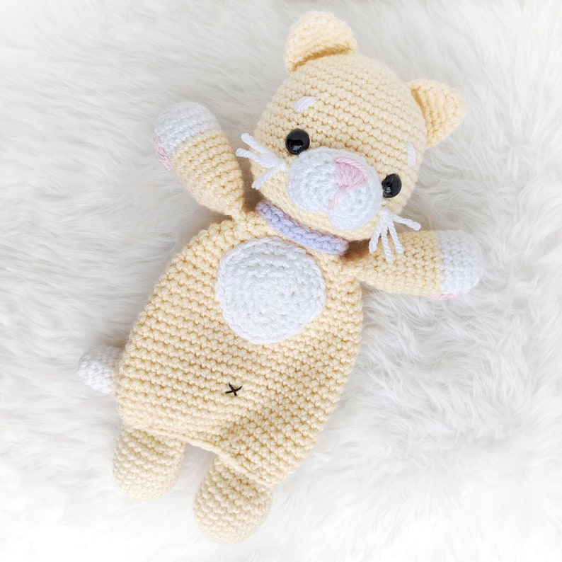 May include: A crocheted stuffed cat toy with a cream-colored body, white accents, and black eyes. The cat is lying on a white fluffy surface.