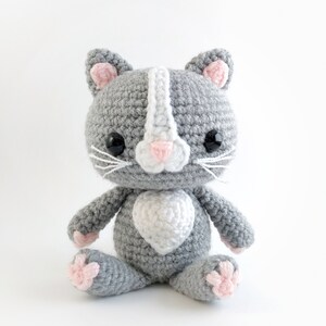 May include: A gray crocheted cat with pink ears and a white heart on its chest. The cat has black eyes and white whiskers.