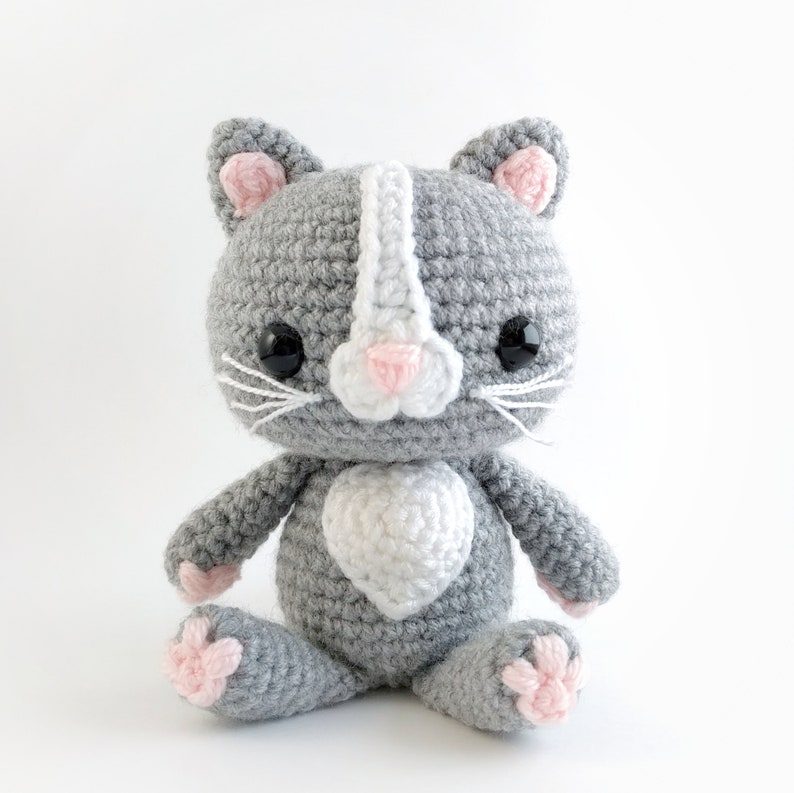 May include: A gray crocheted cat with pink ears and a white heart on its chest. The cat has black eyes and white whiskers.