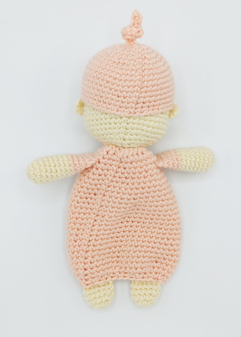 May include: A crocheted baby doll with a pink hat and a pink and cream outfit. The doll is lying on its back with its arms outstretched.