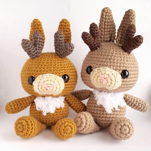 Puede incluir: Dos juguetes de reno de crochet, uno marrón y otro amarillo mostaza, con ribete de piel blanca y ojos negros. Ambos renos tienen cuernos marrones.