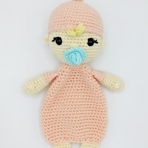 May include: A crocheted baby doll with a pink hat, yellow hair, and a blue pacifier. The doll is wearing a pink crocheted outfit.