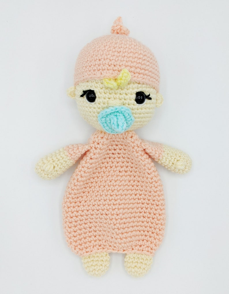 May include: A crocheted baby doll with a pink hat, yellow hair, and a blue pacifier. The doll is wearing a pink crocheted outfit.