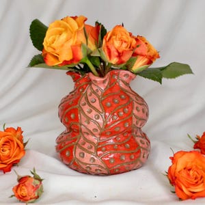 May include: A ceramic vase with a red and pink leaf pattern holds orange roses. The vase has a unique shape and is decorated with a repeating pattern of leaves and dots. The roses are in full bloom, with vibrant orange petals.