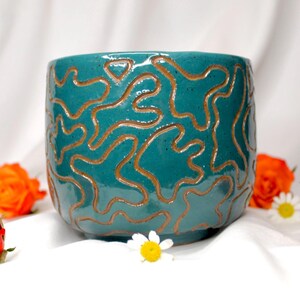 Op de afbeelding: Een turquoise keramische plantenbak met een bruin, abstract, verhoogd ontwerp. De ronde pot heeft een glanzende afwerking en is omgeven door oranje rozen en witte madeliefjes. De plantenbak is geschikt voor binnen of buiten.