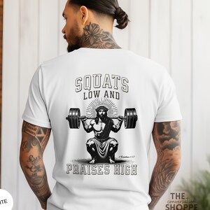 May include: White t-shirt with a graphic of Jesus doing a squat with a barbell. The text on the shirt reads "SQUATS LOW AND PRAISES HIGH" with a reference to 2 Corinthians 1:3-7.
