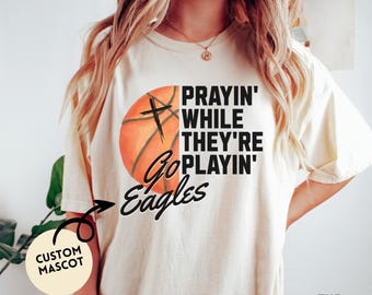 May include: Cream-colored t-shirt featuring a basketball design with a cross and the words "Prayin' While They're Playin' Go Eagles." The shirt has a custom mascot design and is made of soft material.