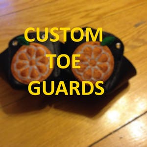 Puede incluir: Un par de protectores de dedos negros con diseños de frutas bordadas en naranja. El texto "CUSTOM TOE GUARDS" está escrito en letras amarillas.