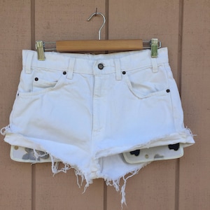 Puede incluir: Pantalones cortos de mezclilla blanca con bordes deshilachados y un forro de estampado floral. Los pantalones cortos están colgados en una percha de madera.