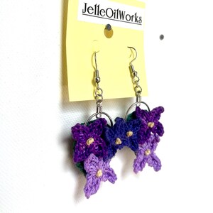 May include: Crocheted flower earrings in shades of purple and lavender with silver-tone ear wires. The earrings are attached to a yellow tag with the text "JelleOilWorks". The flowers have small yellow centers.
