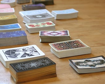 May include: Stacks of tarot cards with various designs, including floral, celestial, and geometric patterns. The cards are arranged on a wooden surface, showcasing the diverse artwork on the card backs and faces. The decks vary in color and design.