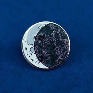 May include: A silver-toned enamel pin featuring a black and white crescent moon design. The moon is depicted with a detailed surface texture and craters.