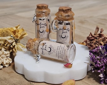 May include: Three small glass jars with cork stoppers and silver charms. Each jar is filled with crystals and has a label that reads "Creativity Jar" with a crescent moon and two hands reaching up. The label also includes the text "A jar to spark inspiration, awaken new ideas, and unleash boundless creativity."