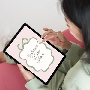 May include: A digital tablet displaying the text "Confidence Begins Inside" within a decorative frame. The frame and bow are a soft green color, set against a pale pink background. The tablet is held by a person wearing a green jacket.