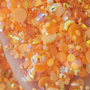 May include: A mix of orange, iridescent, and clear resin embellishments. The embellishments are in various shapes, including circles, ovals, and diamonds.