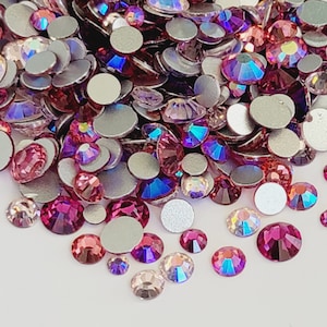 May include: A large assortment of pink, purple, and clear iridescent rhinestones. The rhinestones are scattered on a white surface.
