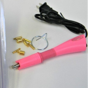 Puede incluir: Una herramienta aplicadora de pedrer&iacute;a rosa con un cable de alimentaci&oacute;n negro, un anillo de metal plateado y varias puntas de pedrer&iacute;a doradas. La herramienta tiene un mango rosa y un agarre rojo.