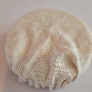 May include: A white cotton fabric cover for a bowl or plate. The cover is round and has a gathered edge.