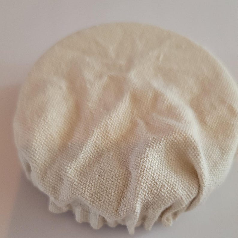 May include: A white cotton fabric cover for a bowl or plate. The cover is round and has a gathered edge.