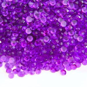 May include: A large pile of purple, round, iridescent rhinestones. The rhinestones are scattered on a white surface.