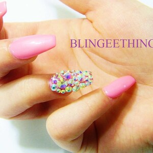 May include: Pink long acrylic nails with a single nail featuring a cluster of iridescent rhinestones.  BLINGEEETHINGEE is written in purple text on the background.
