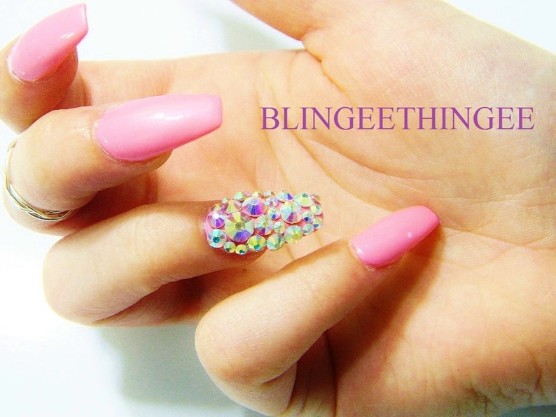 May include: Pink long acrylic nails with a single nail featuring a cluster of iridescent rhinestones.  BLINGEEETHINGEE is written in purple text on the background.