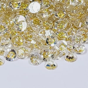 May include: A pile of small, clear, iridescent glitter with gold flecks. The glitter is scattered on a white surface.