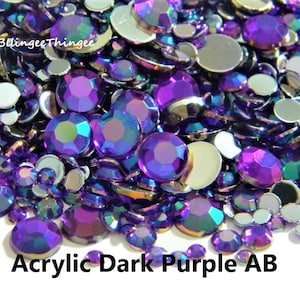 May include: A close-up of a pile of dark purple iridescent acrylic rhinestones. The rhinestones are mixed with silver flatback rhinestones.