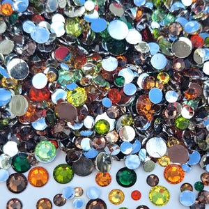 May include: A close-up of a collection of colourful, round rhinestones. The rhinestones are in various colours, including silver, brown, orange, green, yellow, and red. The rhinestones are scattered across a white surface, creating a sparkling effect.