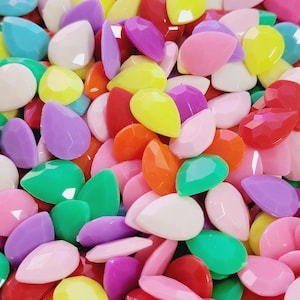 May include: A large assortment of colorful teardrop-shaped rhinestones. The rhinestones are made of plastic and have a glossy finish. They come in a variety of colors, including pink, purple, yellow, green, orange, and white.