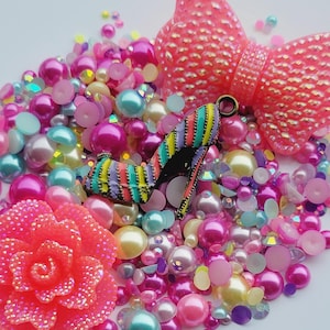 May include: A collection of colorful beads and embellishments, including pink and white pearls, iridescent blue and green beads, and pink and white glitter accents. There is also a pink flower, a pink bow, and a colorful striped shoe charm.