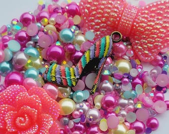 May include: A collection of colorful beads and embellishments, including pink and white pearls, iridescent blue and green beads, and pink and white glitter accents. There is also a pink flower, a pink bow, and a colorful striped shoe charm.