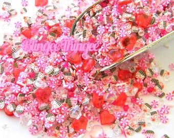Pode incluir: Uma mistura de granulados e enfeites decorativos rosa, vermelhos e brancos, incluindo formatos de coração, doces e cristais transparentes. O texto "Blingee Thingee" é visível. Ideal para artesanato ou decoração.