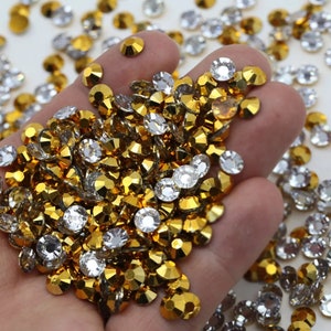 May include: A close-up of a hand holding a large quantity of gold and silver rhinestones. The rhinestones are scattered on a white surface.