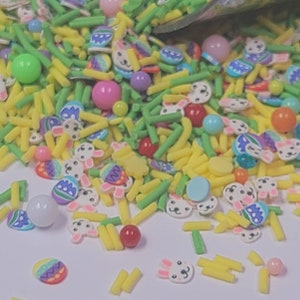 Può includere: Una varietà di zuccherini colorati, tra cui giallo, verde e rosa, a forma di coniglietti, uova e bastoncini. Gli zuccherini sono sparsi su una superficie bianca.