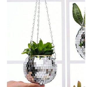 Peut inclure: Un pot de fleurs suspendu sphérique recouvert de petits carreaux de miroir argentés réfléchissants, ressemblant à une boule à facettes. Le pot est suspendu par trois chaînes en métal argenté et contient une plante verte aux feuilles pointues.