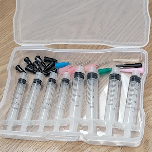 May include: A clear plastic case containing several syringes and needles. The syringes are white with black markings indicating measurements in milliliters. The needles have various colored caps and tips. The case is open, revealing the contents.