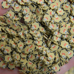 May include: A large pile of small, brown teddy bear shaped resin charms with red bows. The charms are scattered on a hand.