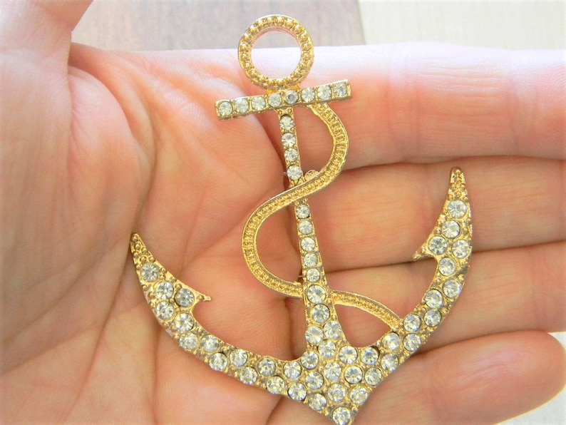 May include: A gold-tone anchor brooch with clear rhinestones. The anchor is detailed with a rope design and has a classic shape.