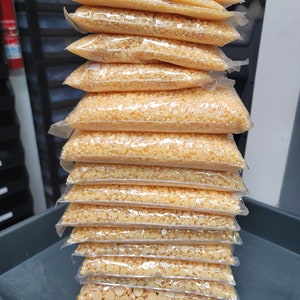 Könnte beinhalten: Ein Stapel klarer Plastiktüten, gefüllt mit kleinen, hellorangen, runden Perlen. Die Beutel sind versiegelt und ordentlich aufeinander gestapelt. Die Perlen scheinen gleichmäßig groß und geformt zu sein, und die Beutel sind in einer hohen Säule angeordnet.