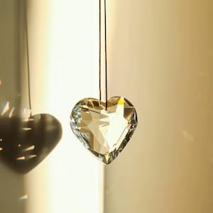 May include: A crystal heart-shaped ornament hangs from a brown string. The ornament is clear and reflects light.