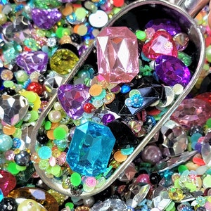 May include: A silver scoop filled with a variety of colorful rhinestones, gems, and beads. The scoop is sitting on a bed of similar colorful rhinestones, gems, and beads.