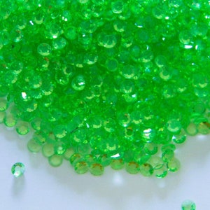 May include: A large pile of bright green, round, faceted rhinestones. The rhinestones are scattered on a white surface.