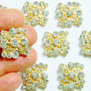 May include: A close-up of ten gold-toned, square-shaped embellishments with clear rhinestones. The embellishments are scattered on a white surface.