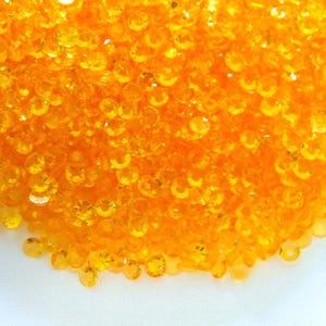 May include: A close-up of a pile of small, round, orange-colored sequins. The sequins are scattered on a white surface.