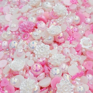 May include: A collection of pink and white pearl-like beads, buttons, and decorative embellishments. The beads and buttons are in various shapes and sizes, including flowers, circles, and ovals.