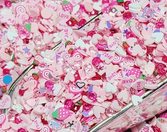 Pode incluir: Uma colher de confeitos de resina rosa, branca e transparente com várias formas, incluindo corações, estrelas, morangos e espirais. Os confeitos têm a palavra "LOVE" escrita neles.