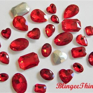 May include: Assortment of red crystal rhinestones in various shapes and sizes, including hearts, ovals, and rectangles.  The rhinestones are scattered on a white surface.