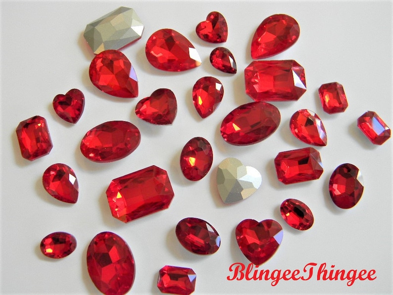 May include: Assortment of red crystal rhinestones in various shapes and sizes, including hearts, ovals, and rectangles.  The rhinestones are scattered on a white surface.
