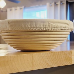 May include: A round, light brown, woven bread proving basket with a white linen liner. The basket is made of natural materials and has a smooth, polished finish.